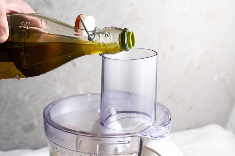A person's hand pouring extra virgin olive oil into a canned sardine spread in a food processor. 