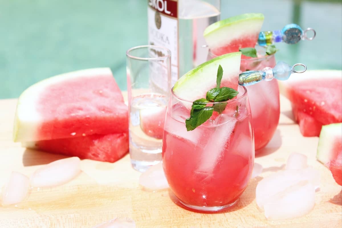 Bottle of vodka, a watermelon drink and slices of watermelon beside a swimming pool.
