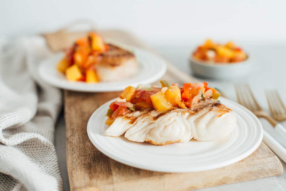 A piece of grilled halibut on a white plate topped with peach jalapeno salsa.