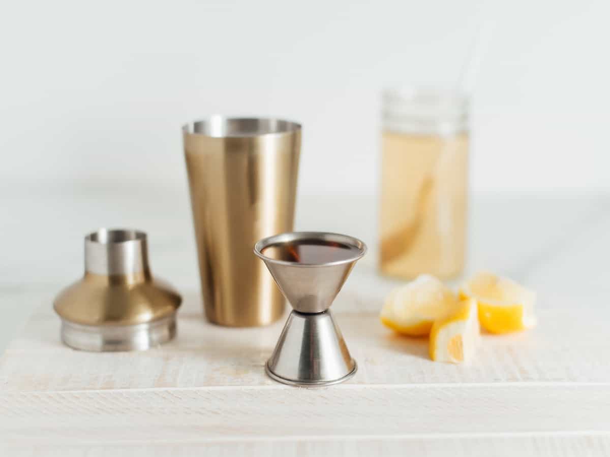 a silver jigger, a drink shaker, lemons, and a clear jar on a light surface