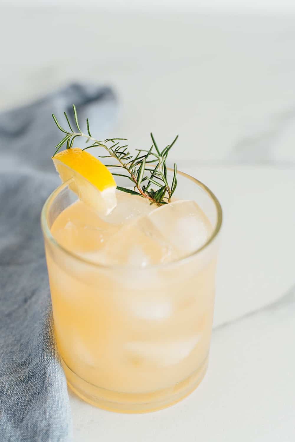 a highball glass, of cahmomile whisky cocktail, filled with ice and liquid with lemon, rosemary, honey syrup in the background