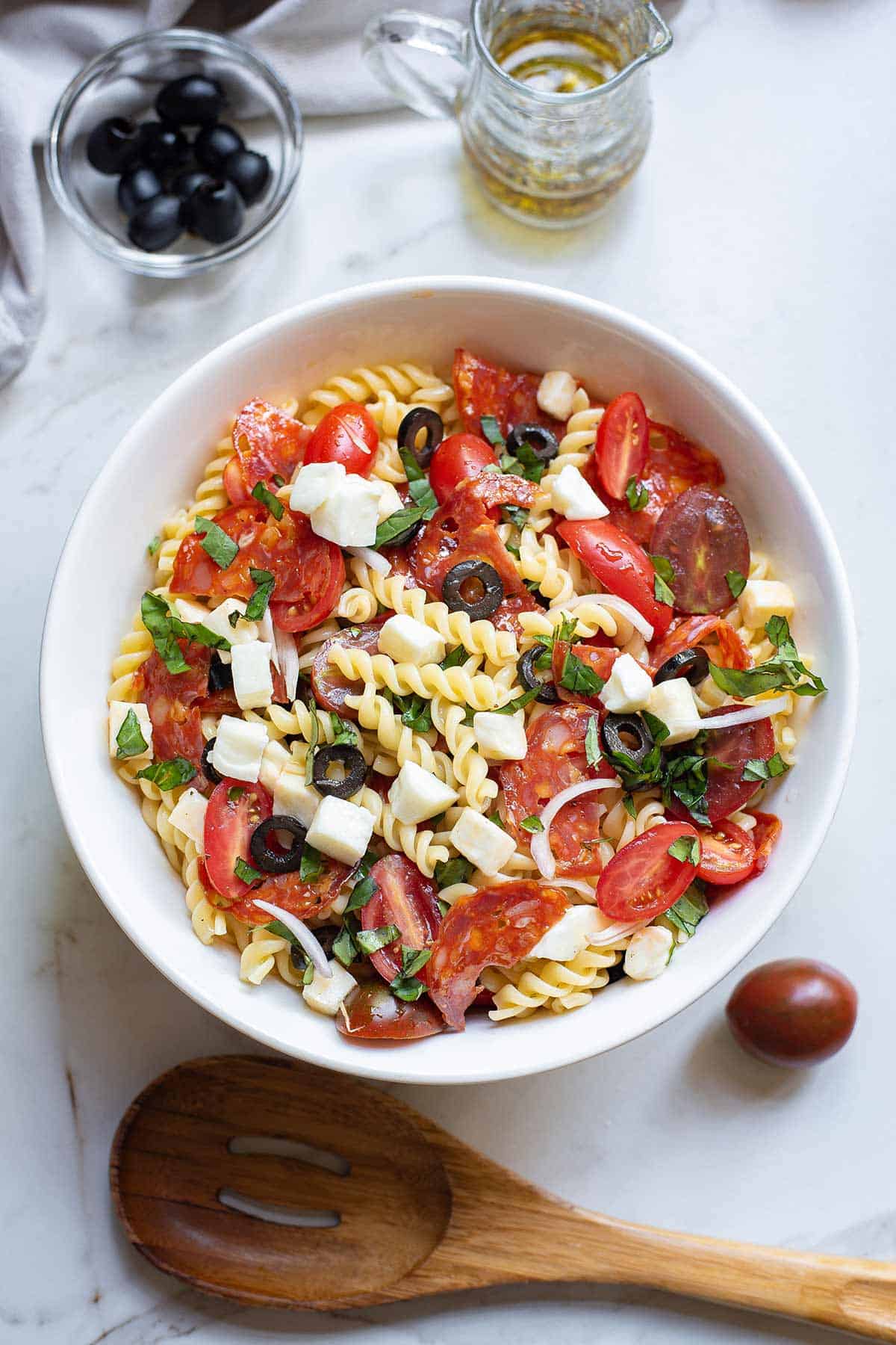 A bowl of pizza pasta salad on the table with a wooden spoon next to the bowl.