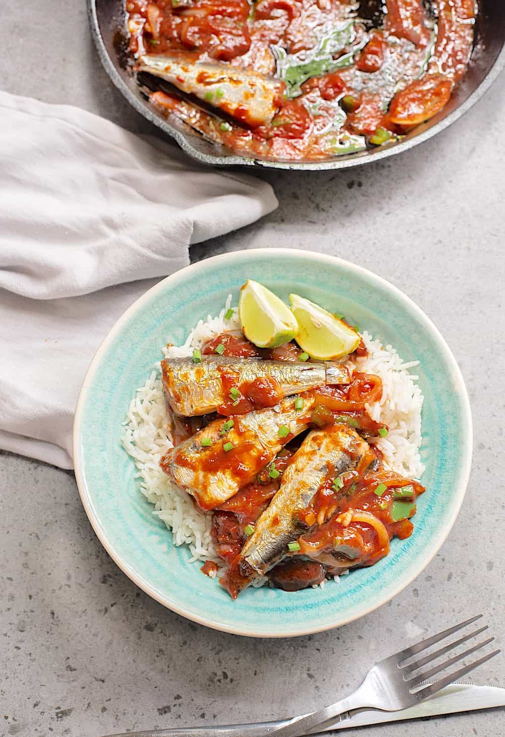 A pan of cooked sardines and a bowl of white rice topped with spicy cooked tomato and sardines.  
